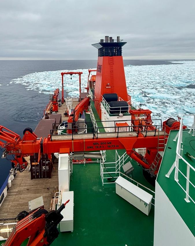 View from the deck of a ship with bright orange cranes and equipment in the foreground, cutting through a field of floating sea ice under a gray, overcast sky.