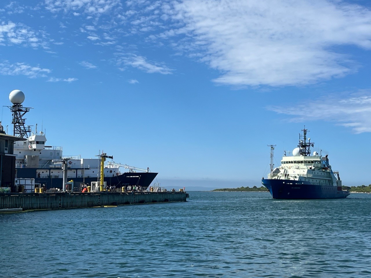 Photo of R/V Neil Armstrong leaving the dock in Woods Hole on a sunny day with bright blue skies. The ship is to the right and the dock is to the left.