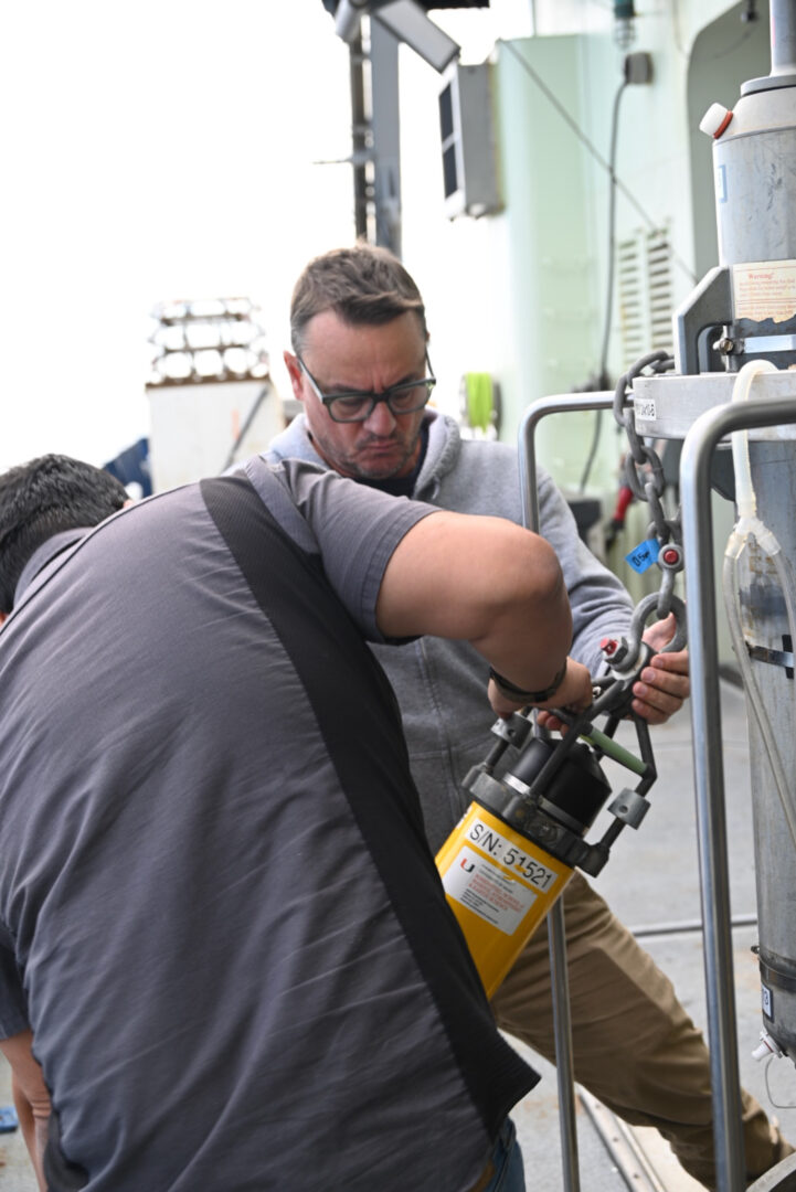 Two people are holding an acoustic release and working together to attach it to the CTD.
