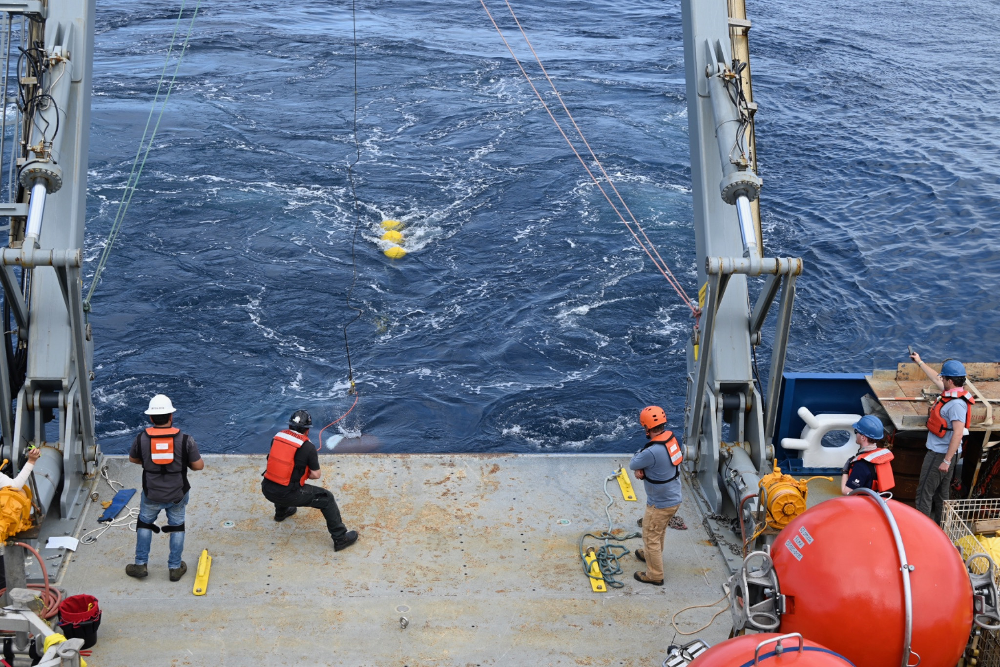 One person releases a line of a mooring into the ocean as four others watch the floatation spheres drift away into the ocean.
