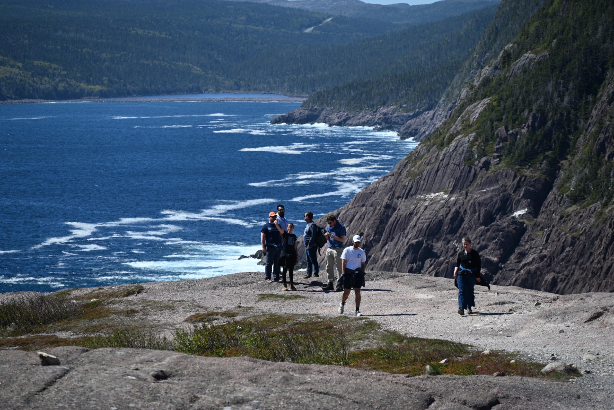 A group of seven people stand on a rocky surface. There are rocky cliffs and bright blue ocean behind them.