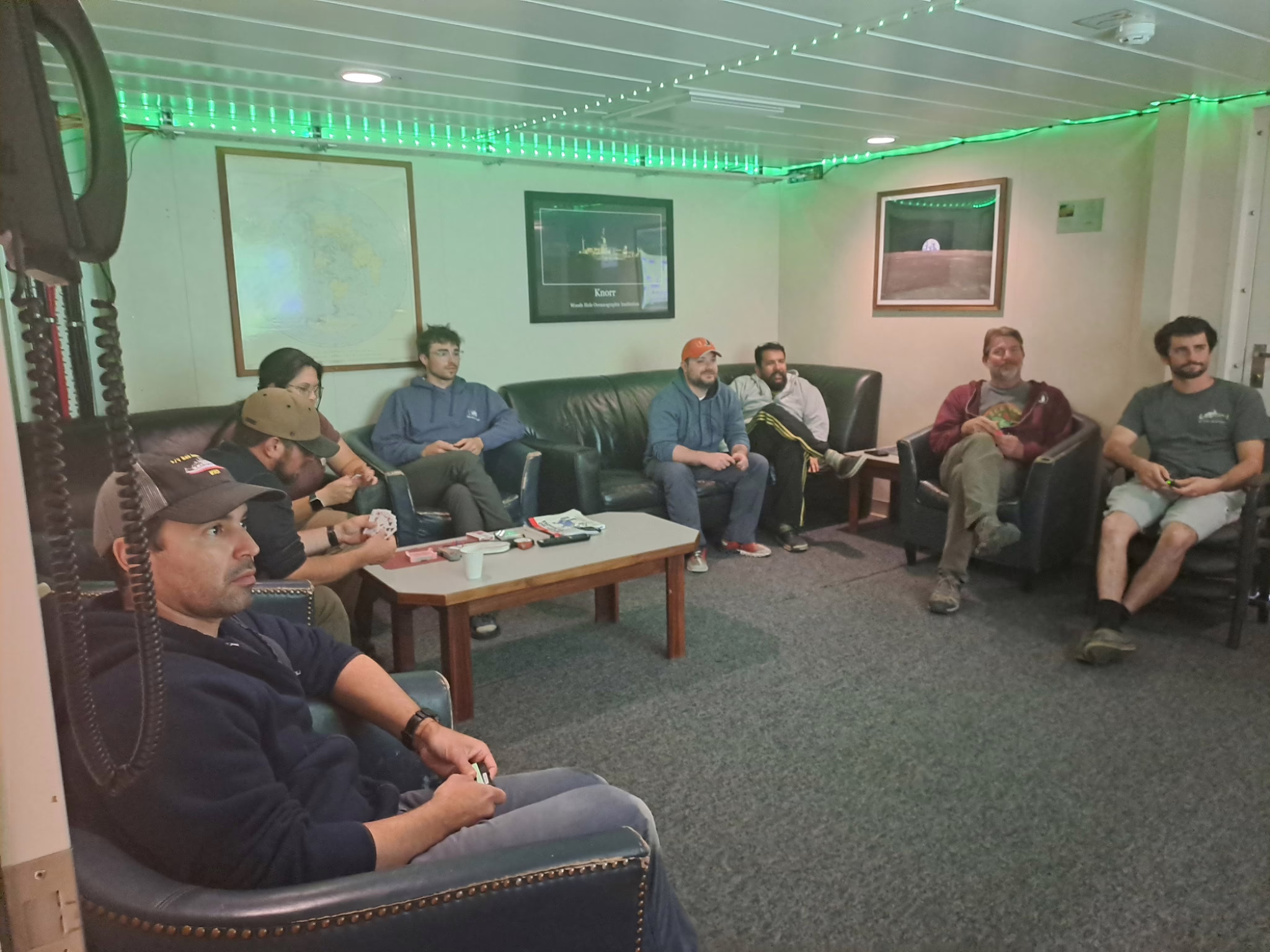 Eight people sit on couches and chairs that surround the room. They appear to be looking at a screen that is not in the photo. A green string of LED lights boards the ceiling.