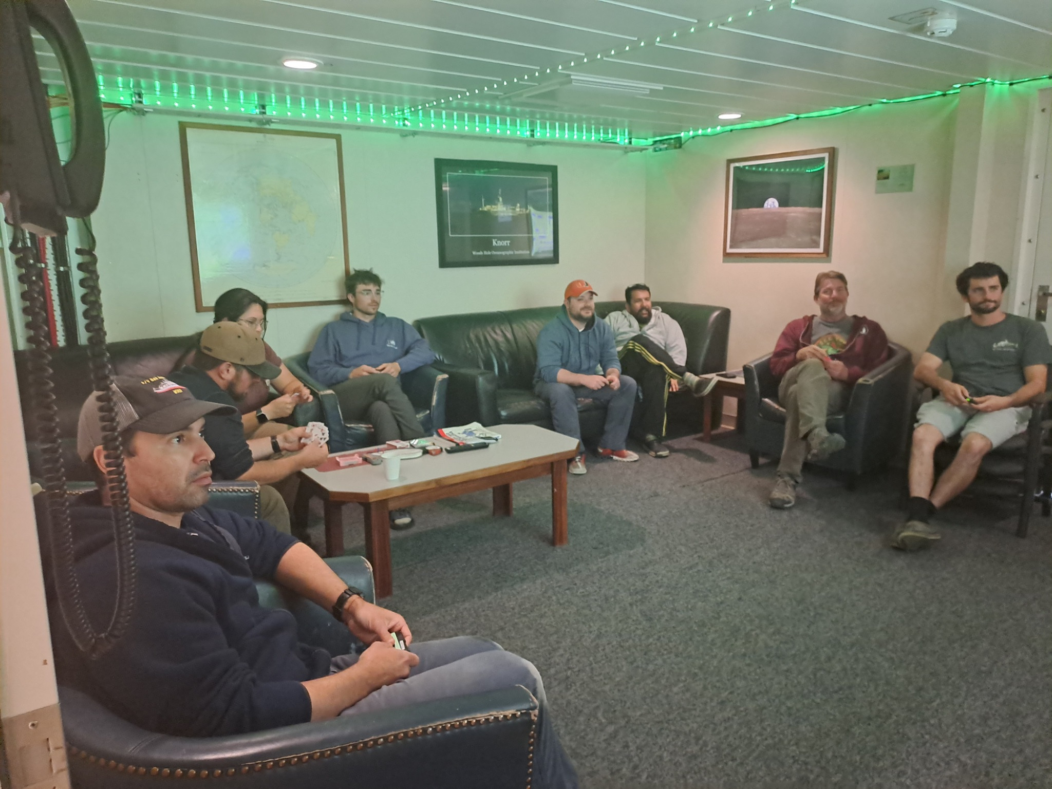 Eight people sit on couches and chairs that surround the room. They appear to be looking at a screen that is not in the photo. A green string of LED lights boards the ceiling.