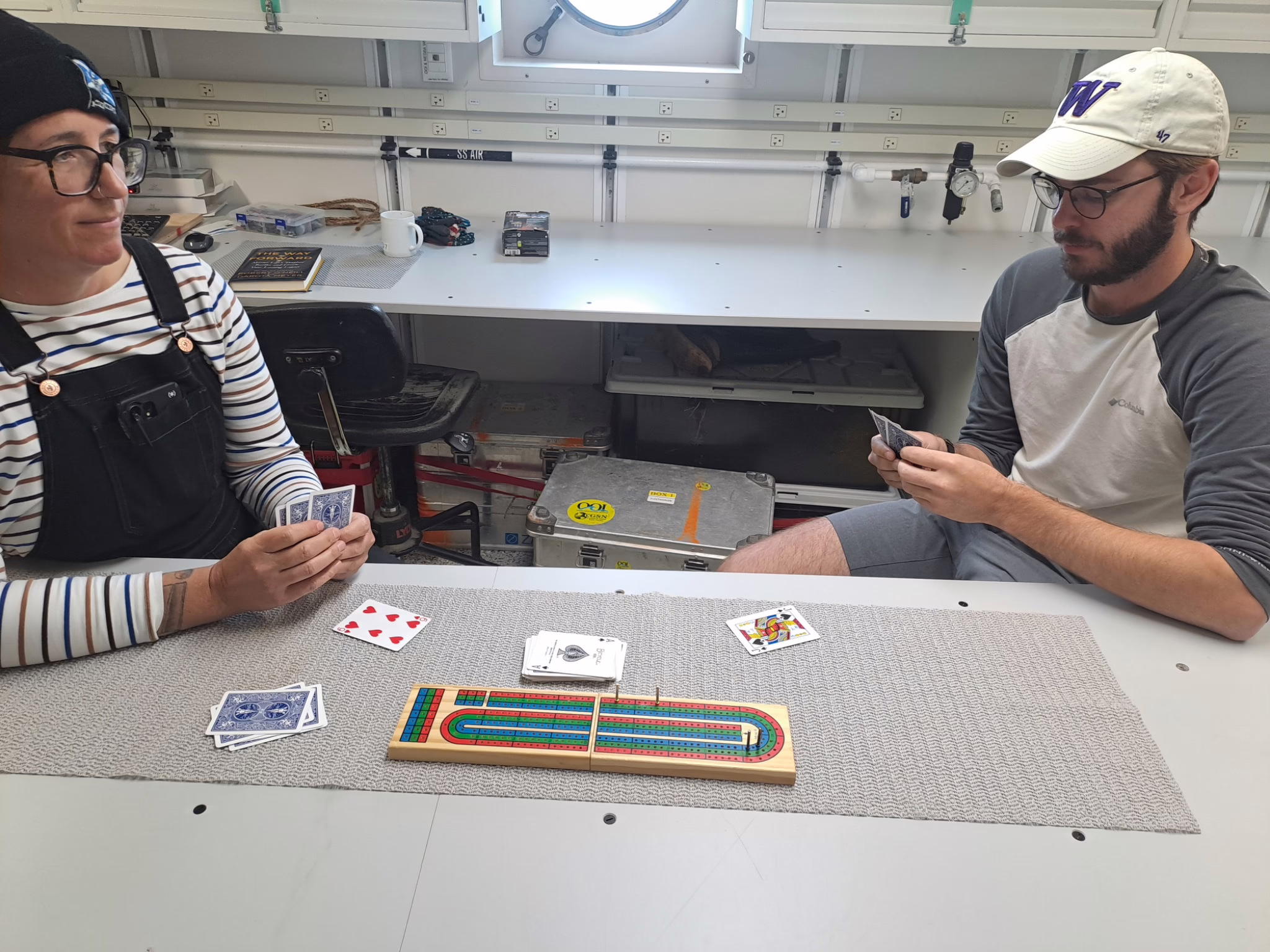 Two people sitting at a lab bench holding and looking at the hand of cards. A cribbage board is between them with some more cards on the table.