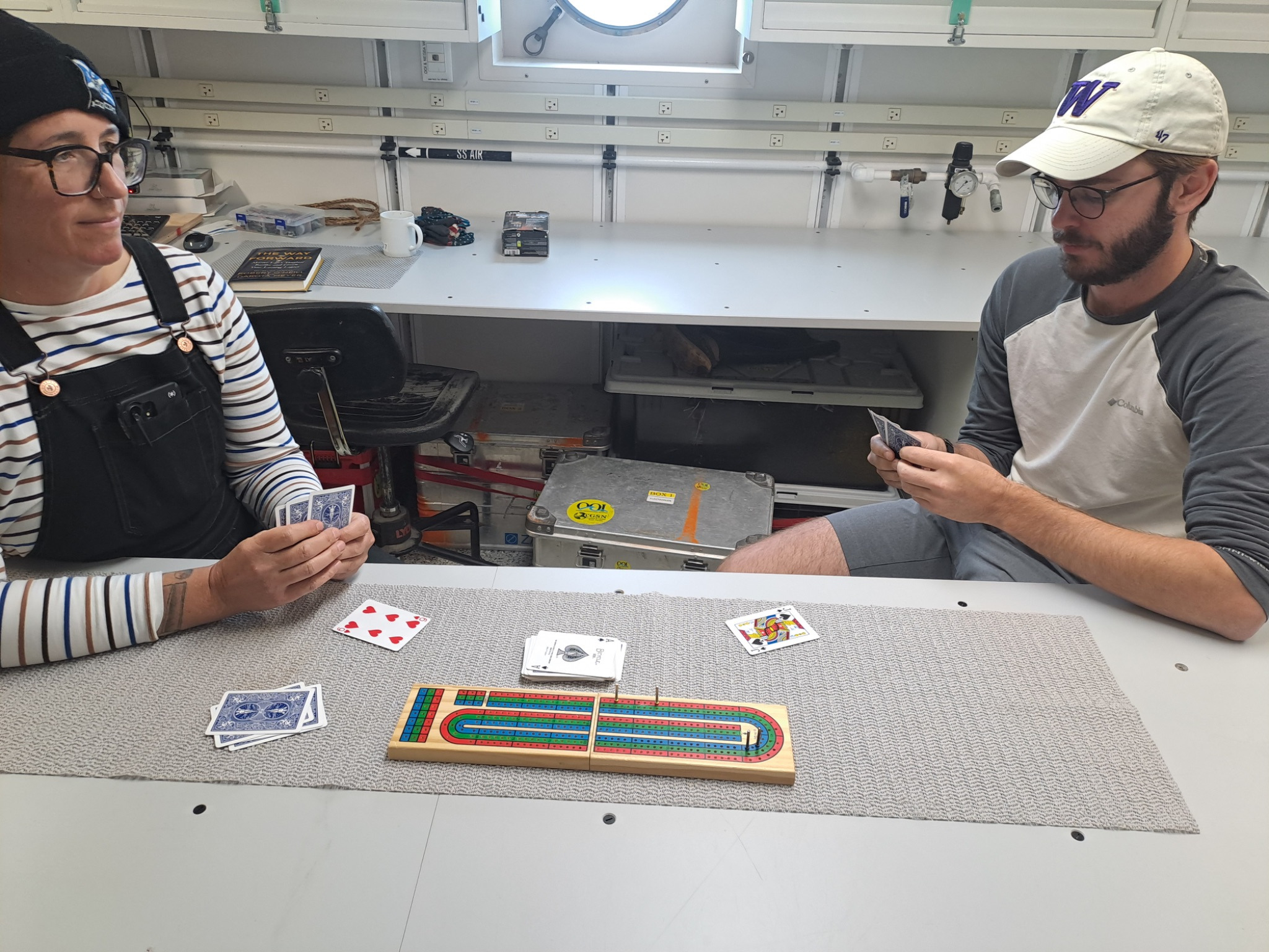 Two people sitting at a lab bench holding and looking at the hand of cards. A cribbage board is between them with some more cards on the table.