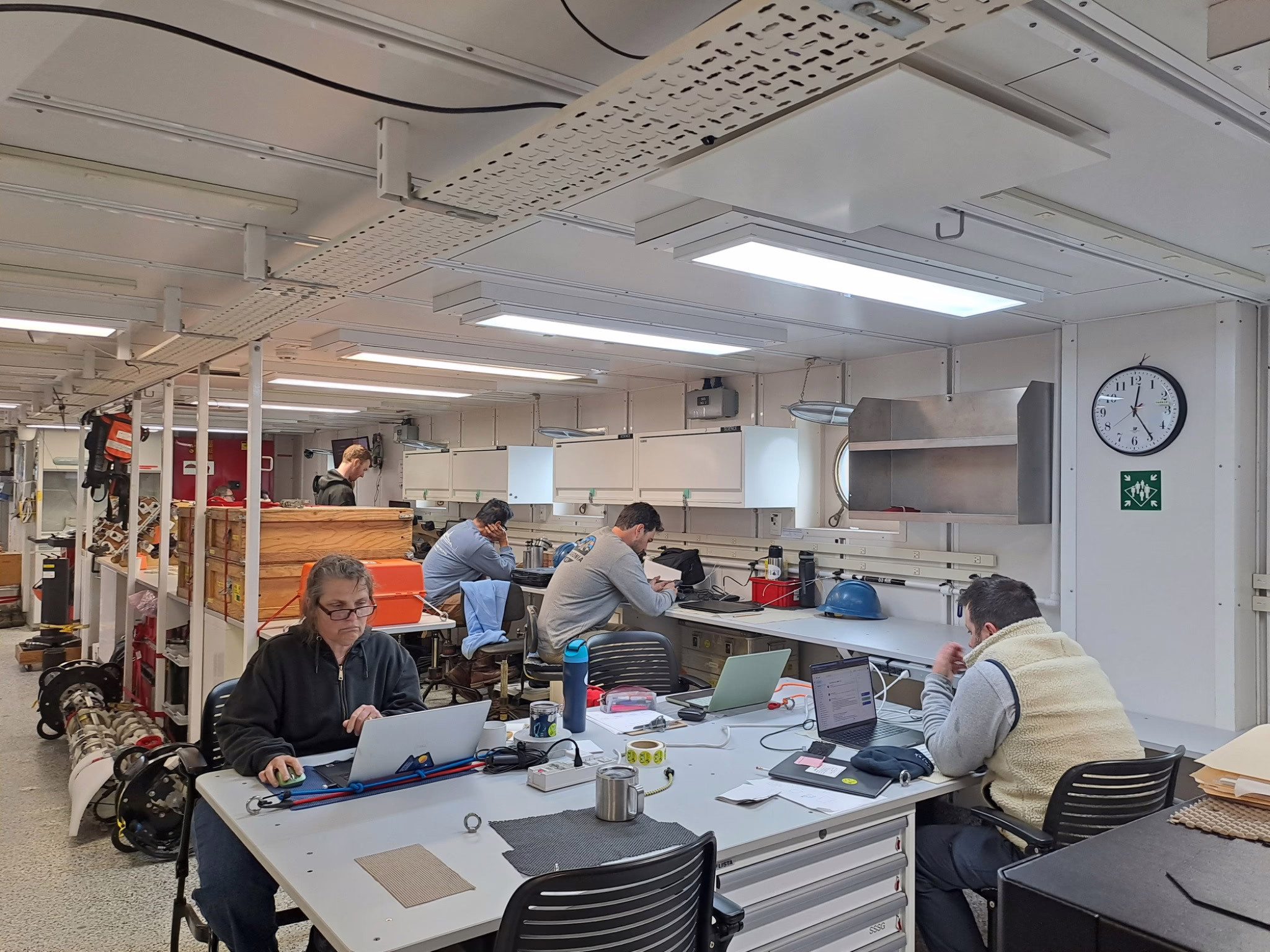 Five people are sitting at work benches and desks looking at their computers and notebooks in the main laboratory aboard the research vessel.