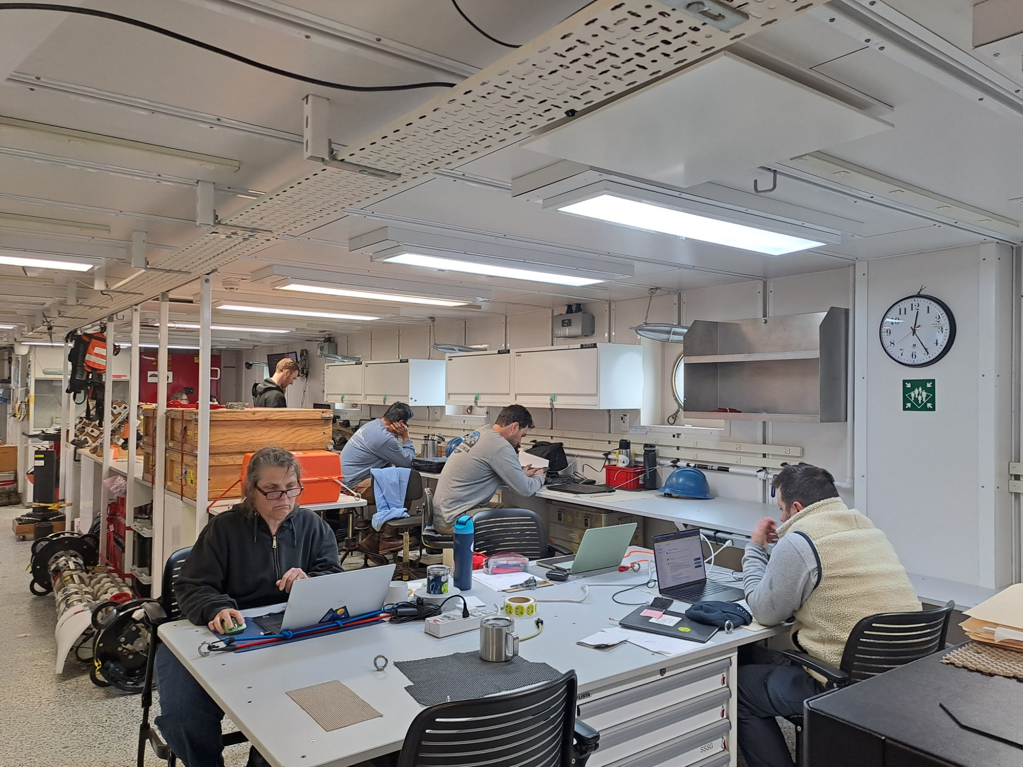 Five people are sitting at work benches and desks looking at their computers and notebooks in the main laboratory aboard the research vessel.