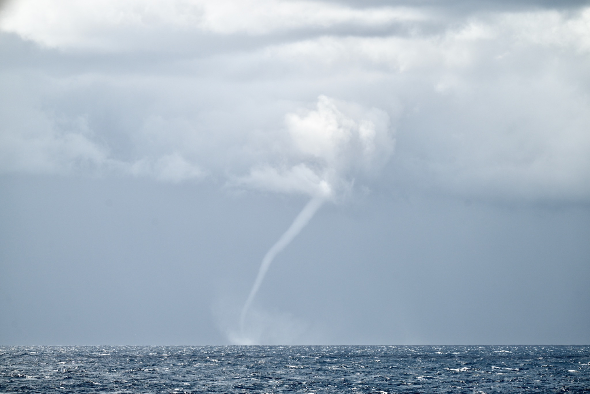 Clouds are seen at the top of the image which turns to fully gray skies below. In the center there's a lighter gray water spout that extends down to the ocean surface. Sort of resembles a tornado in the distance.