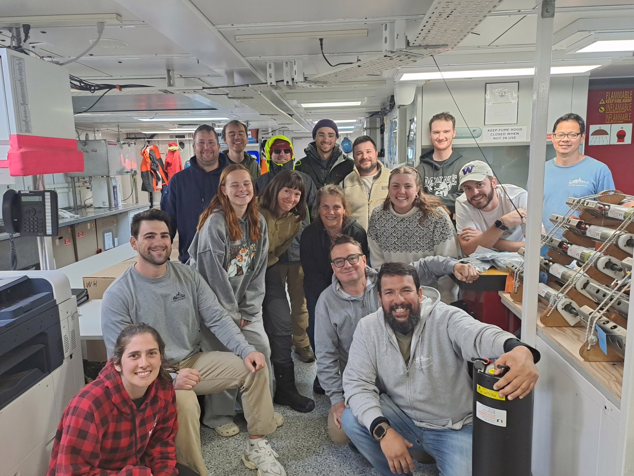 A group of sixteen people gather together and smile for a photo on board the research vessel.