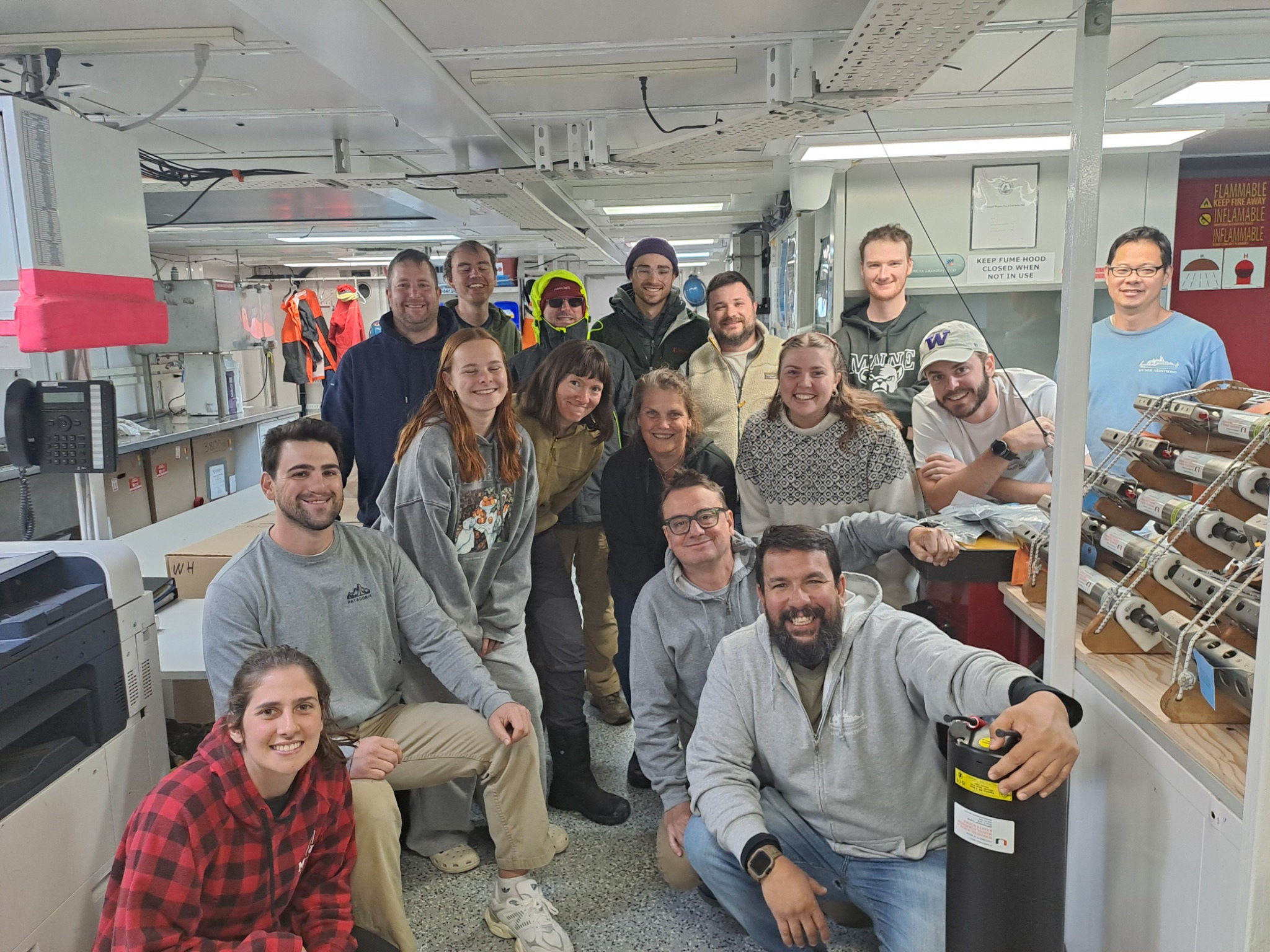 A group of sixteen people gather together and smile for a photo on board the research vessel.
