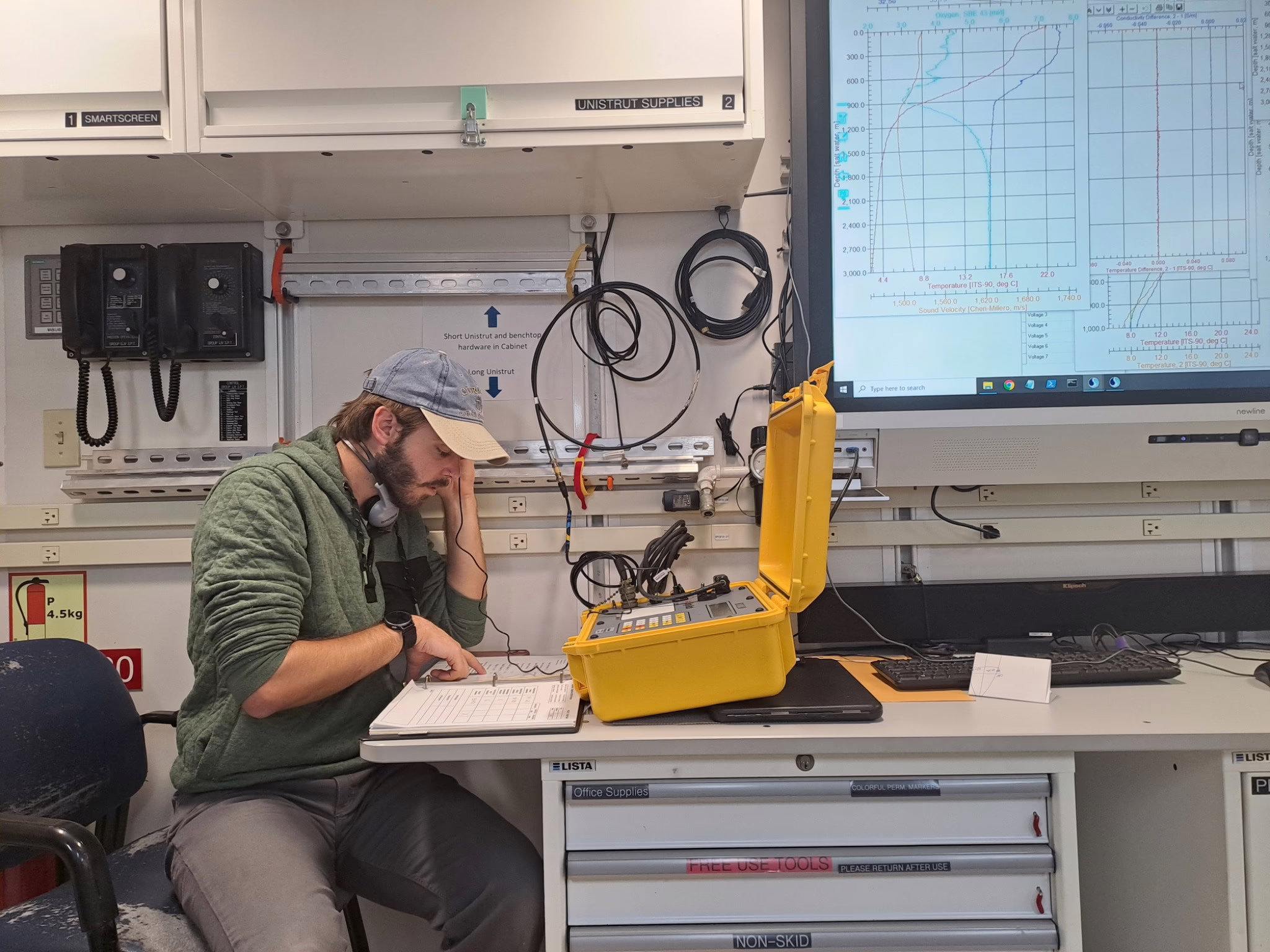 A researcher is sitting at a desk on board a research vessel. He's looking down and pointing at a notebook, has headphones around his neck and scientific equipment is in front of him.