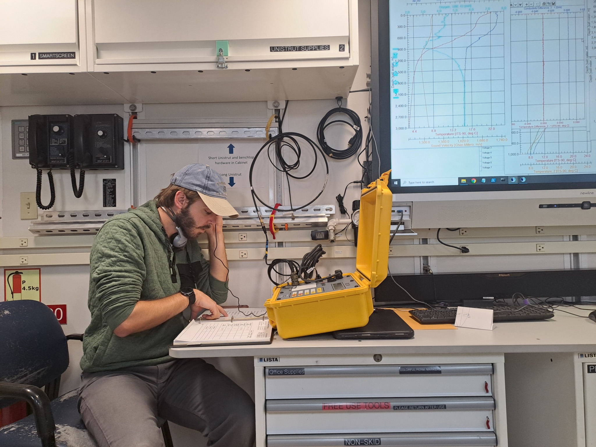 A researcher is sitting at a desk on board a research vessel. He's looking down and pointing at a notebook, has headphones around his neck and scientific equipment is in front of him.