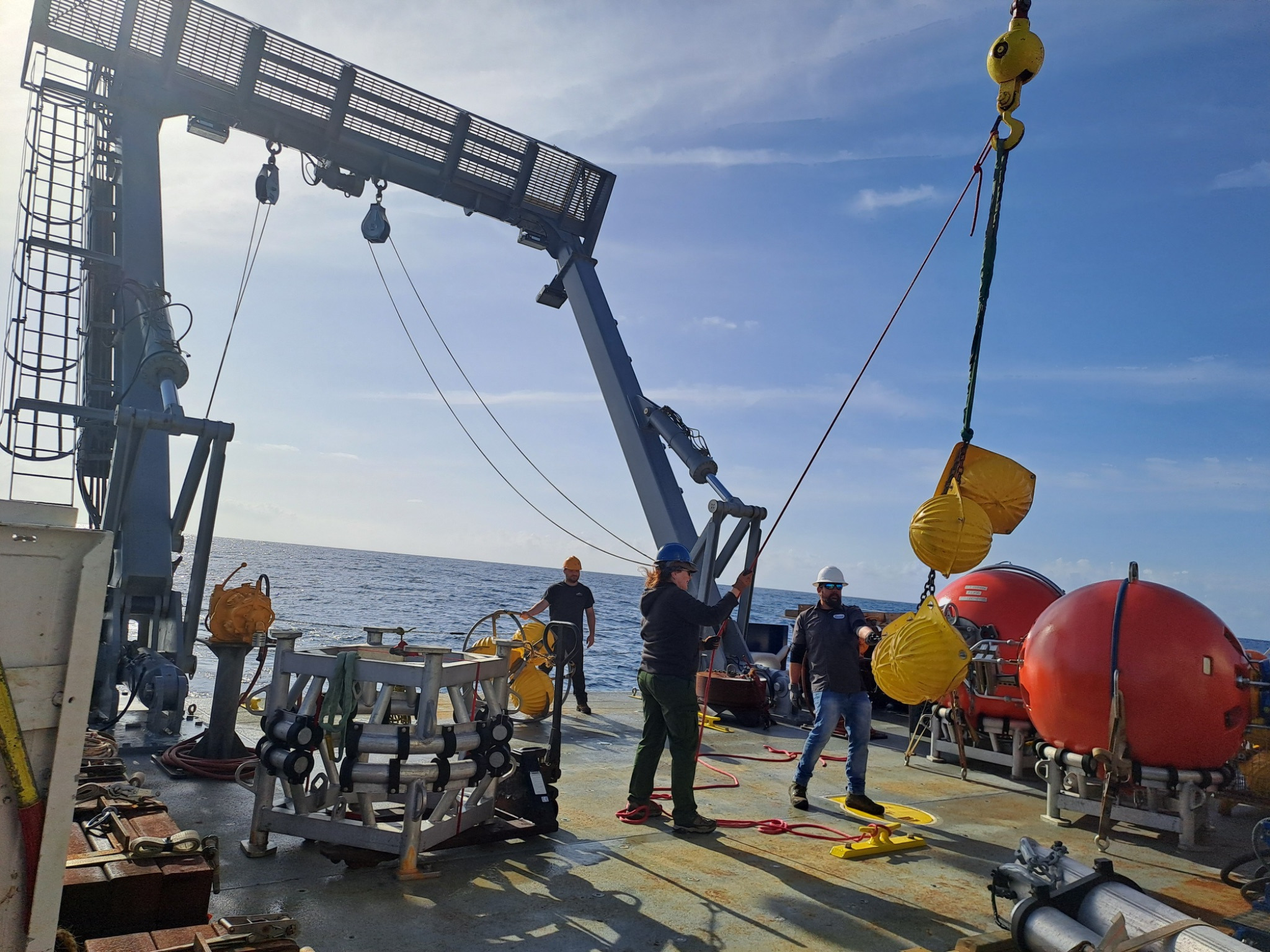 Three people are on the back deck of the ship working together to get a chain with yellow spheres attached to the A-frame. The skies are blue with some wispy clouds.
