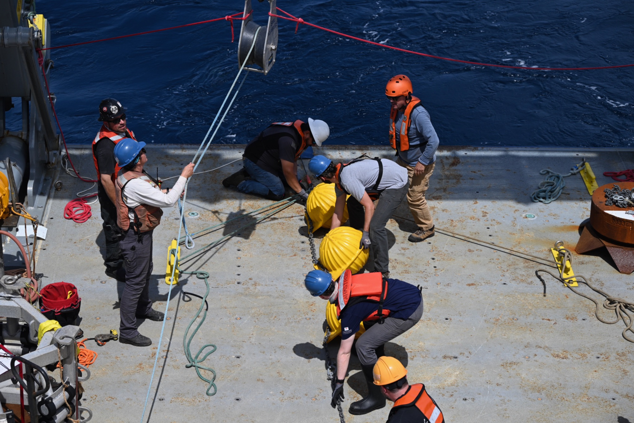 seven people with hard hats and life vests on work together on the back deck to start getting the mooring into the water.