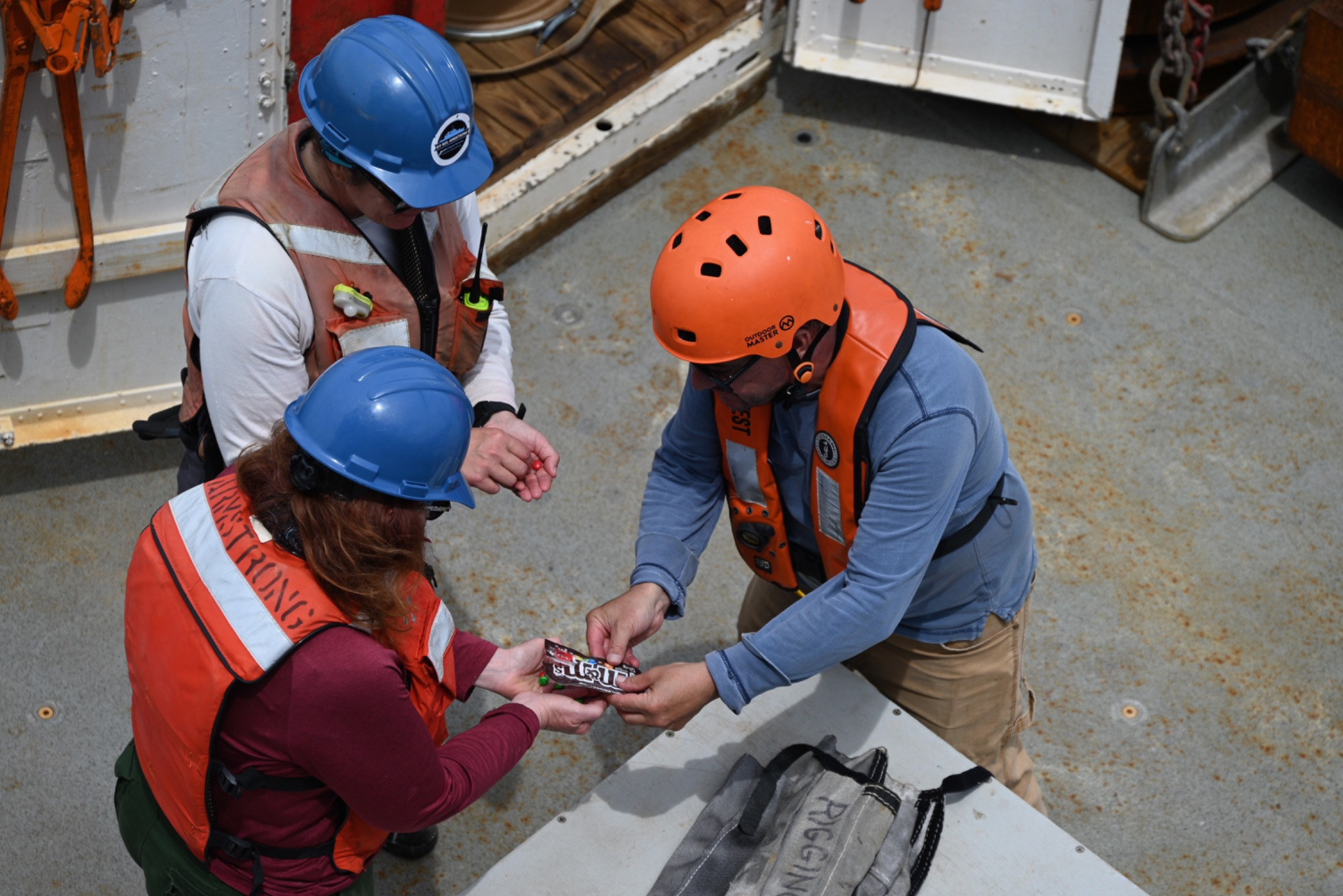 Three people in hard hats and life vests share M&Ms on the deck of a ship.