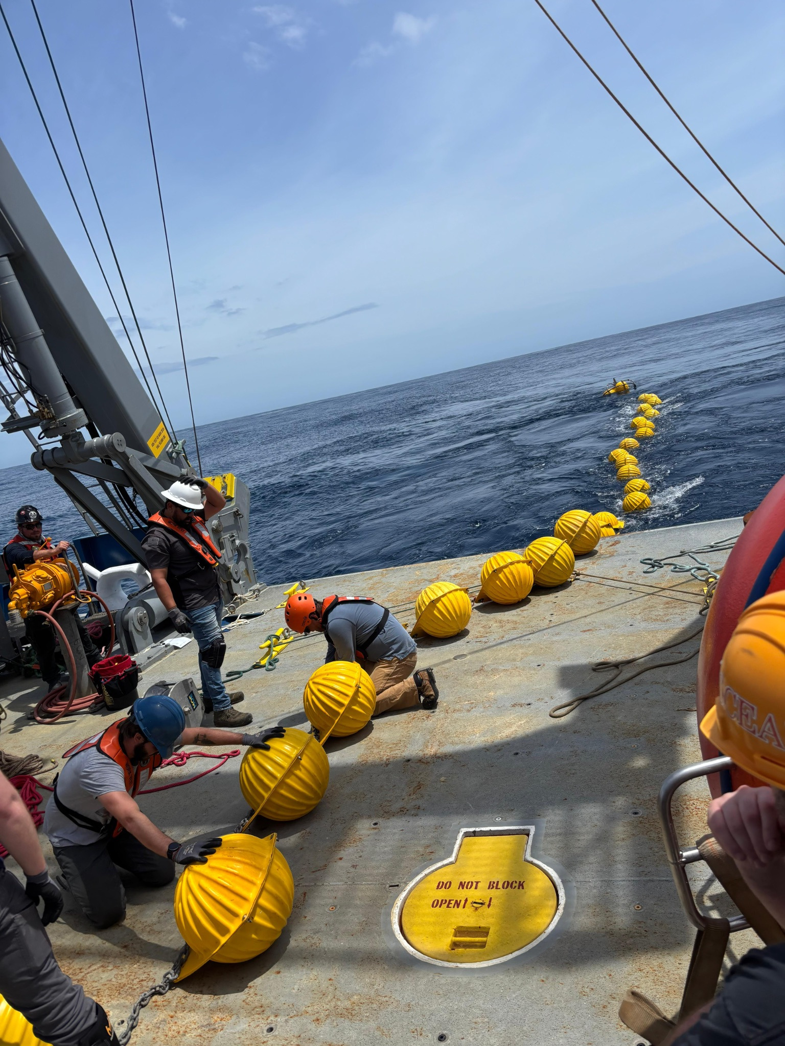 A long line of yellow floatation balls. Half of them are in the ocean, the other half are on the back deck being worked with by a few people in hard hats and life vests.