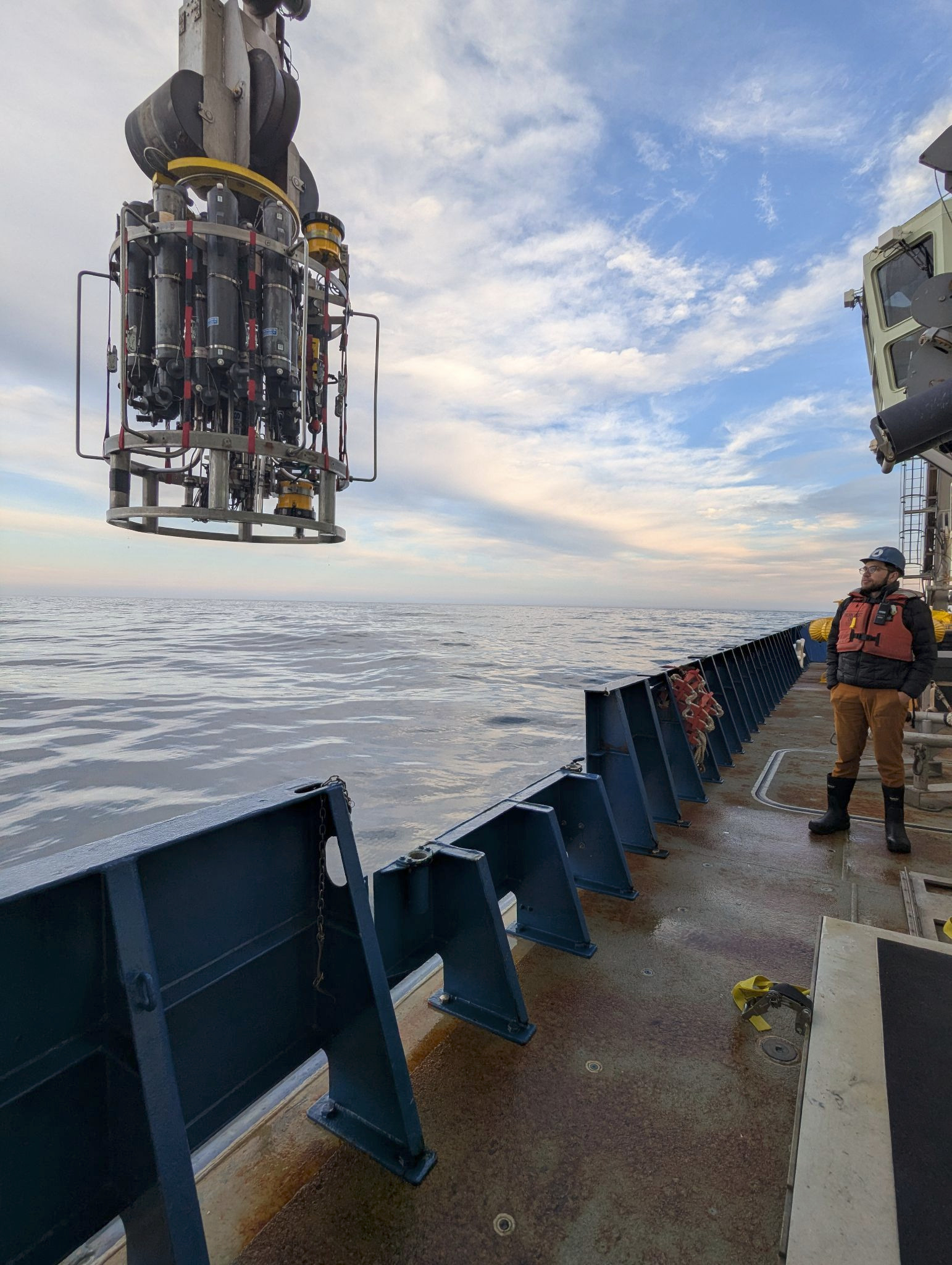 Jed is wearing an orange life vest on the side of the ship. He's watching the CTD rosette being deployed. The seas appear calm with blue and partially cloudy skies.