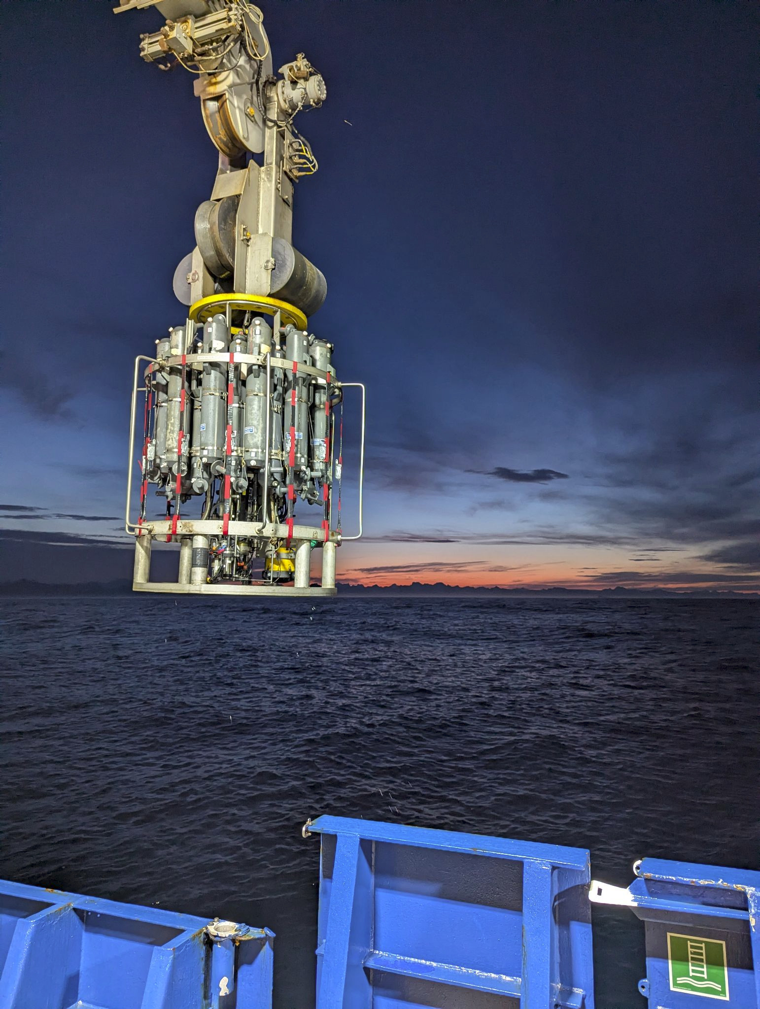 CTD rosette being lowered into the deep, dark, blue sea as the sun is nearly set. There's a speck of orange along the horizon, but mostly deep blue skies with some scattered clouds.