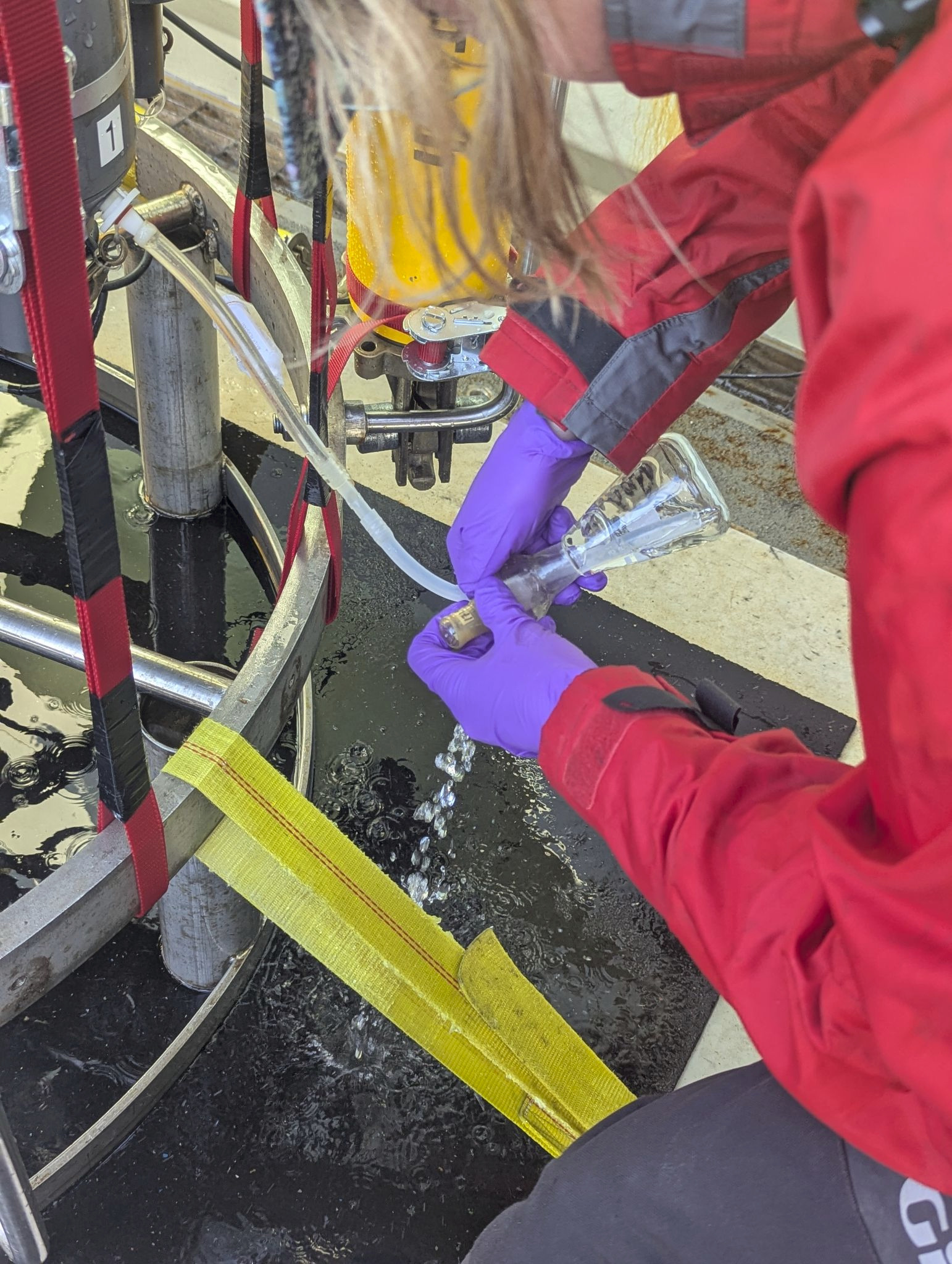 Lucy is wearing a red jacket and purple gloves as she fills flasks with water from the CTD.