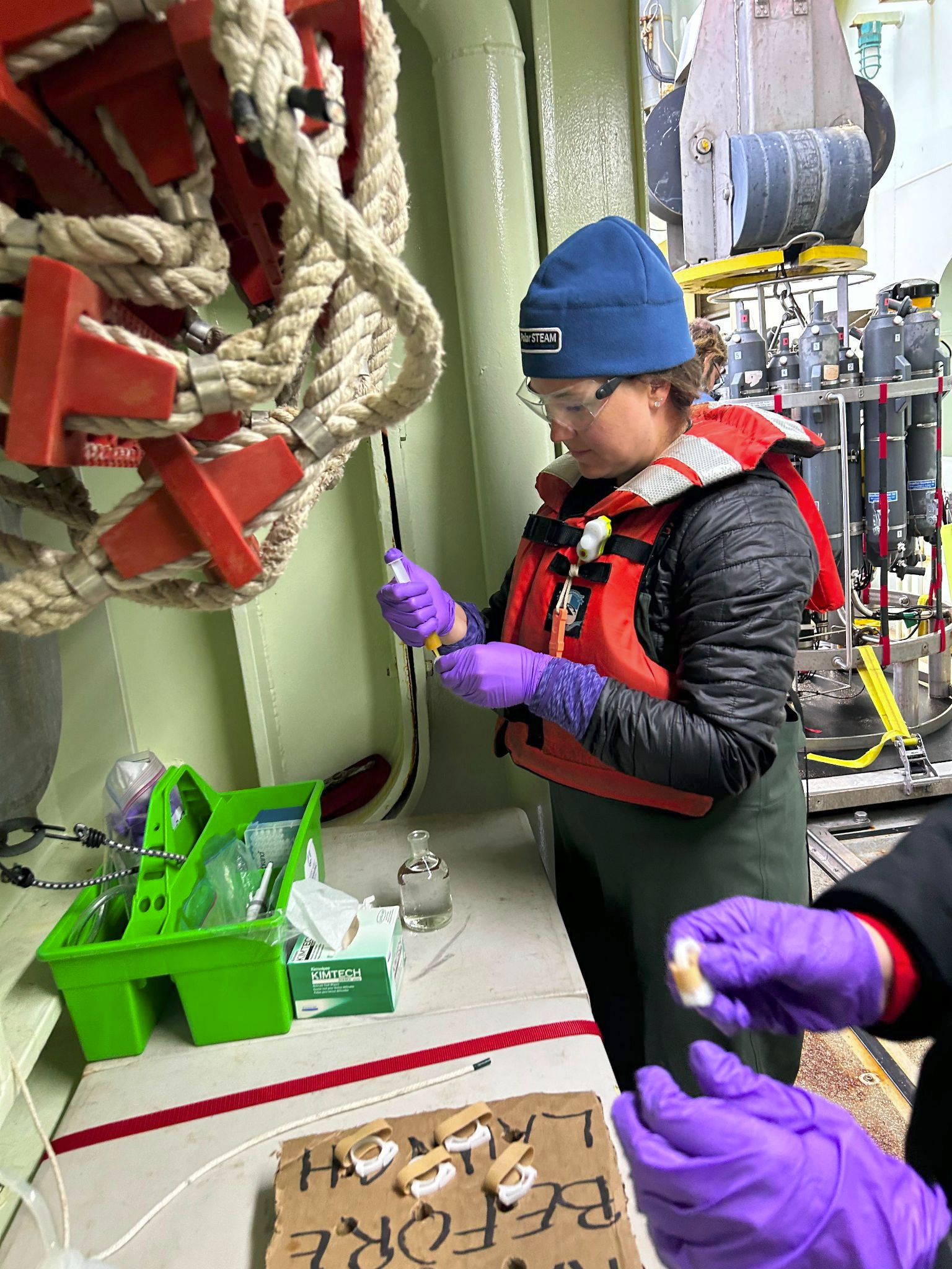 Kathleen is wearing an orange life vest, blue hat, purple gloves and safety goggles as she uses a syringe to poison and grease bottles on the deck of the ship. You can see the CTD rosette behind her.