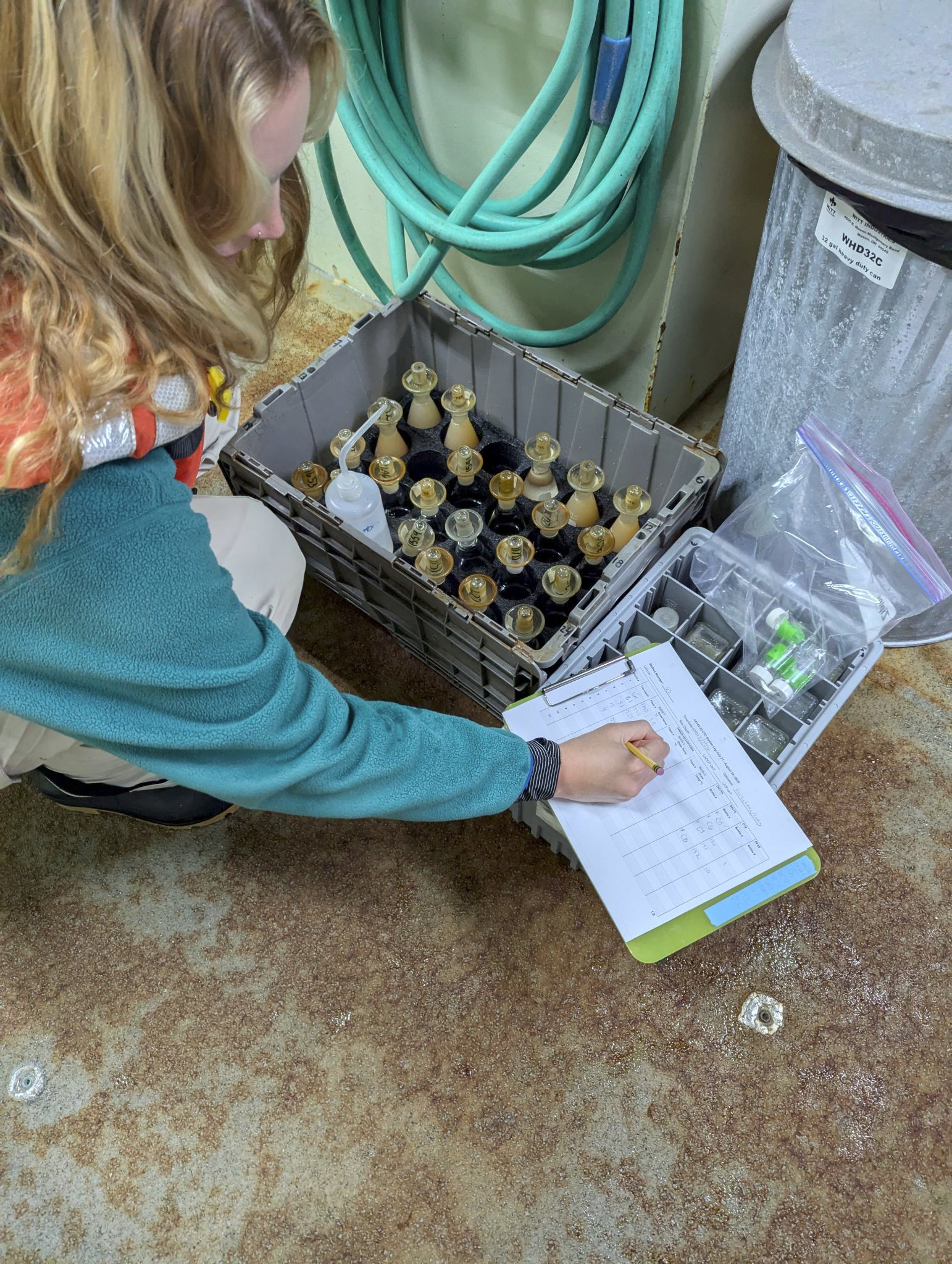 Kylie is knelt down writing on a clipboard next to a bin filled with flasks.