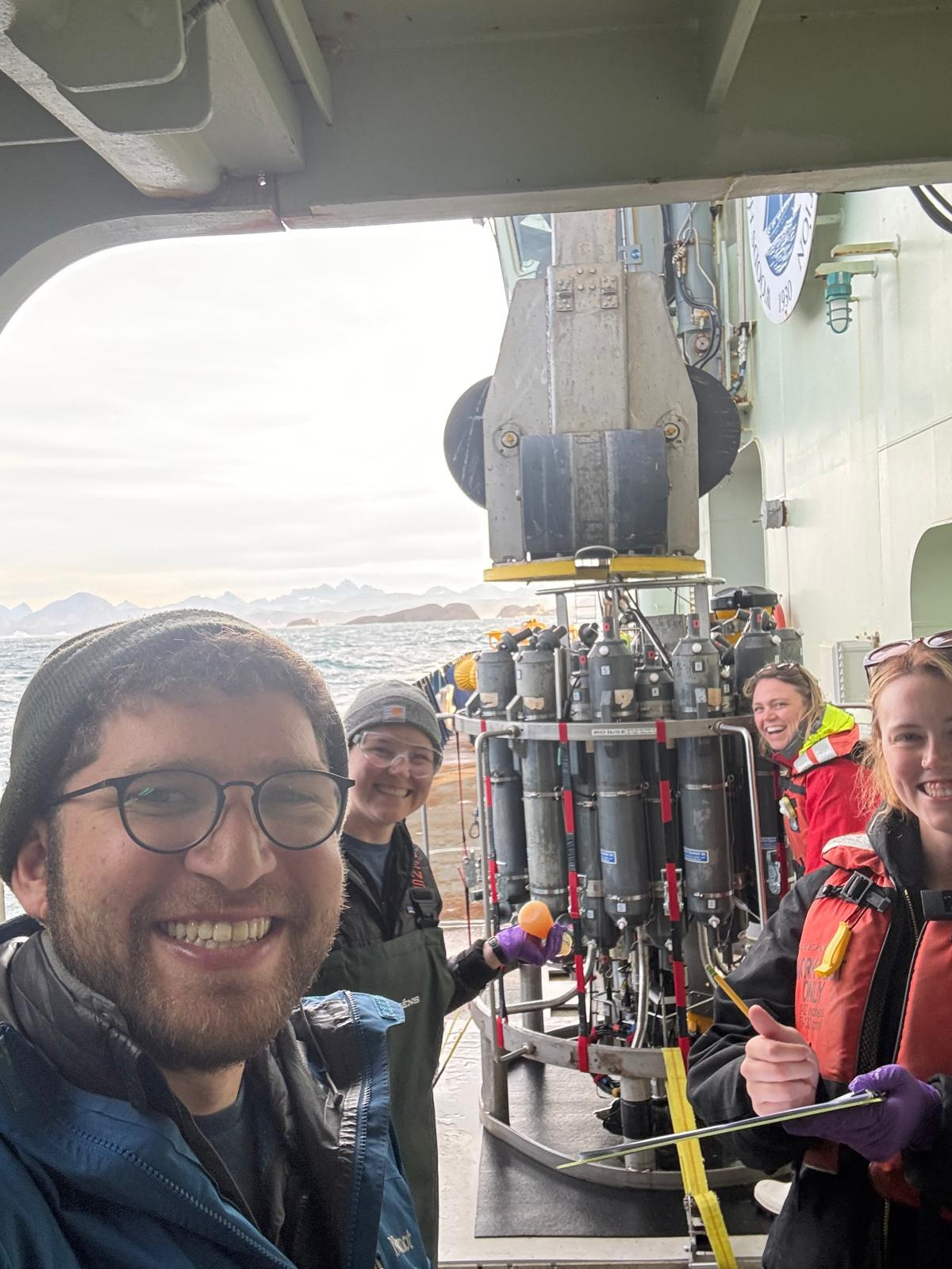 A "selfie" of four people with the CTD in the center of the photo. In the background, you can see the Greenland coastal mountains. Everyone is smiling!