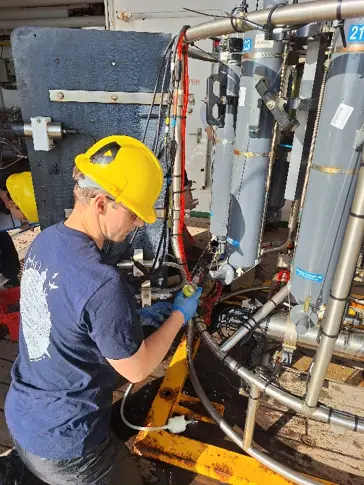 a person in a yellow hard hat and blue gloves taking a water sample from the CTD rosette.