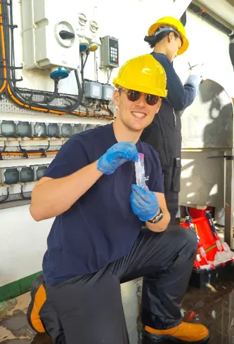 Person in yellow hard hat, sunglasses and blue gloves kneeling on one knee proudly holding a nutrient sample and smiling for the camera.