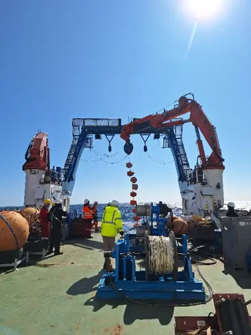 View of the back deck of the ship with at least five people supporting mooring recovery operations. Eight floatation spheres are hanging vertically .