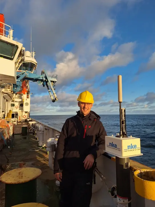 Person in black jacket and yellow hard hat smiles for the camera on the deck of the ship. There are blue skies with some gray patchy clouds.