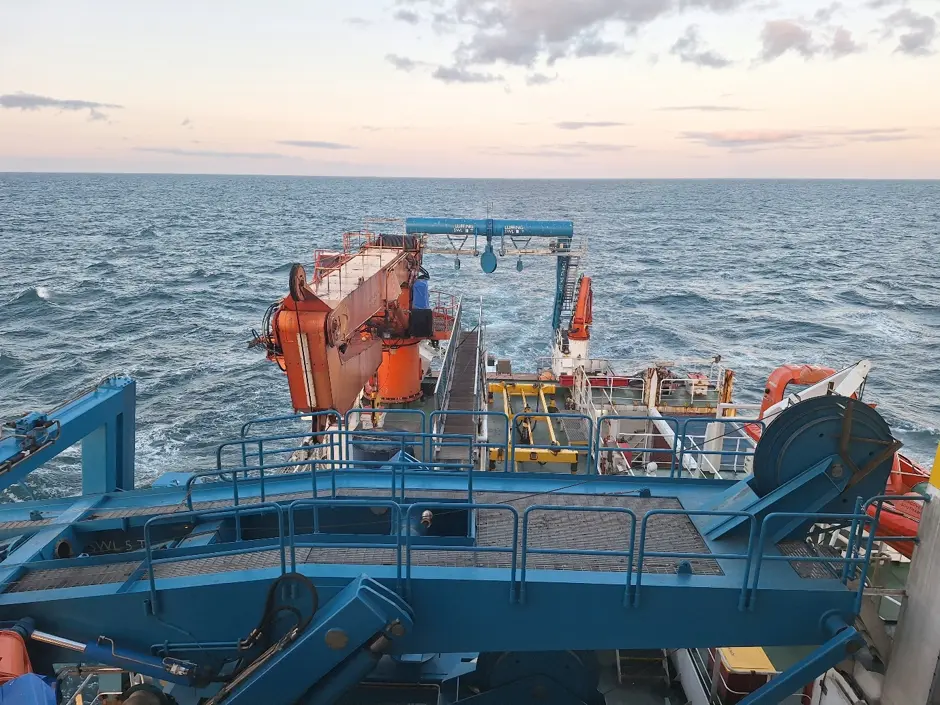 Blue and Orange stern of the RRS Discovery with the deep blue open ocean in the background. The sky has pastel shades of pinks oranges and blues.